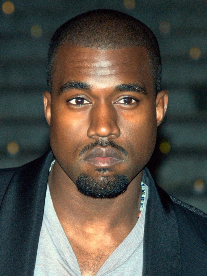 Kanye West mit schwarzen Haaren und schwarzem Bart. Er schaut ernst leicht an der Kamera vorbei.