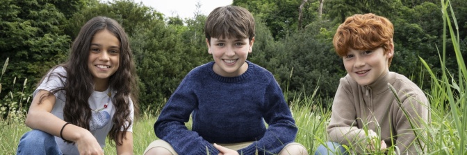 3 Kinder, 2 Jungen und ein Mädchen, knien in einer Wiese und lächeln direkt in die Kamera.