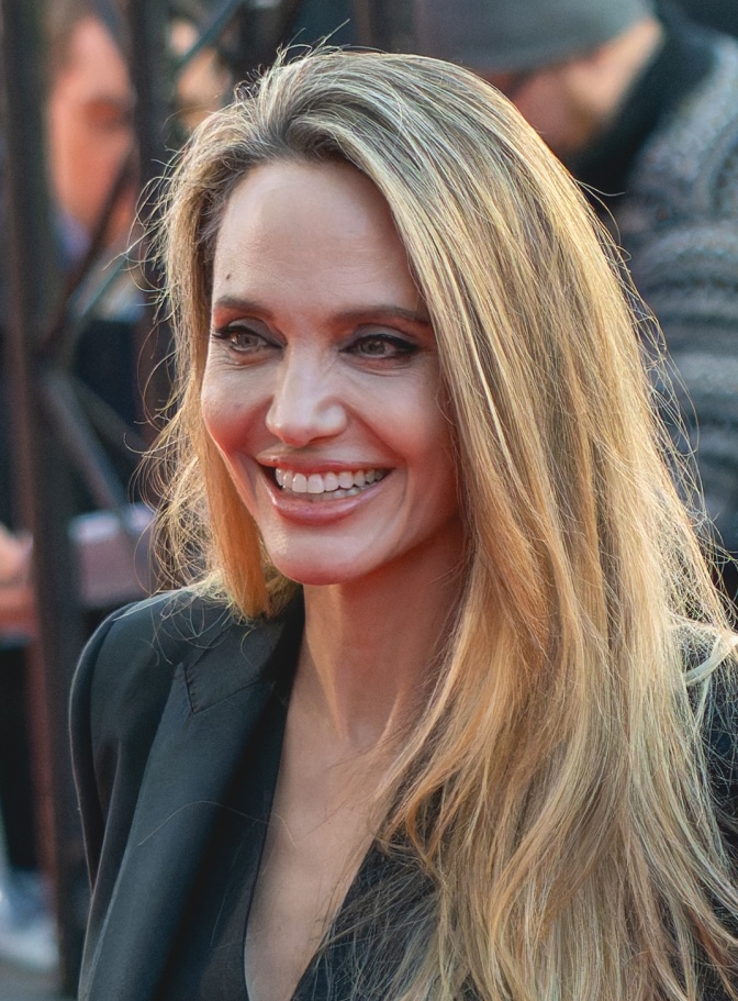 Angelina Jolie mit langen, glatten, gesträhnten Haaren. Sie lächelt direkt in die Kamera, hinter ihr steht eine Menschenmenge.