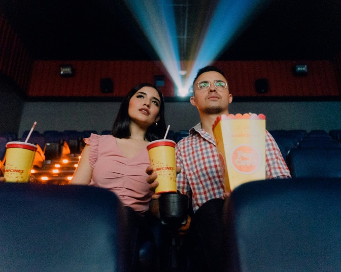 Ein Mann und eine Frau sitzen im abgedunkelten Kinosaal, zwischen sich eine Portion Popcorn. Beide halten einen Getränkebecher in der Hand und schauen nach vorn auf die Leinwand.