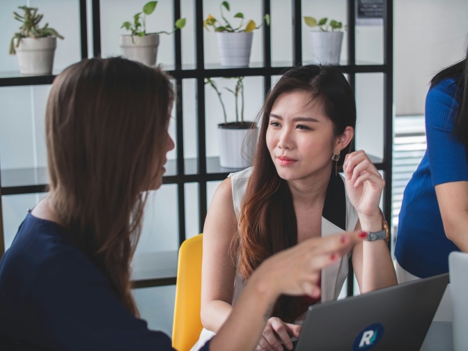 Zwei asiatische Frauen sitzen am Tisch und sind im Gespräch. Eine der beiden schaut die andere skeptisch an.