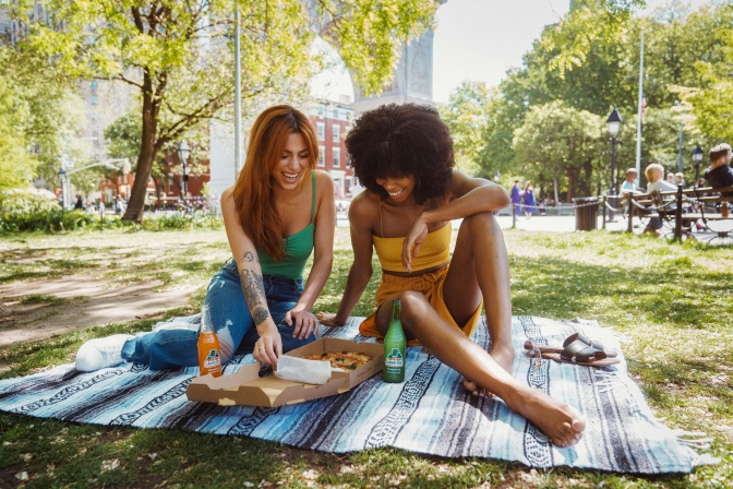 2 Frauen sitzen auf einer Decke und Essen Pizza aus einer Pappschachtel. Eine der beiden Frauen ist weiß, die andere schwarz.