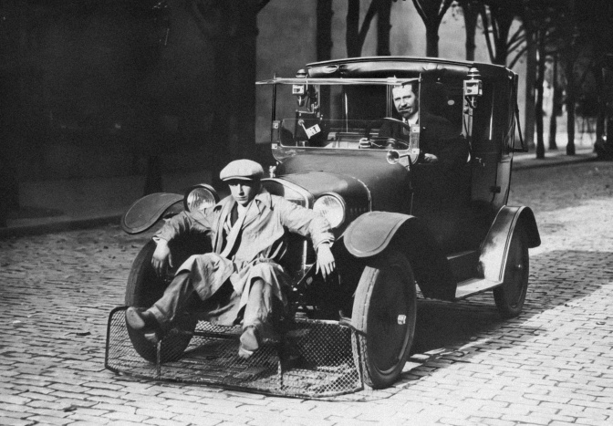 Schwarz-weiß-Foto von 2 Männern, einer von beiden sitzt in einem Auto, der andere hockt davor auf der Stoßstange.