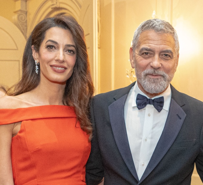 Amal und George Clooney in festlicher Kleidung. Sie stehen nah beieinander und lächeln in die Kamera.