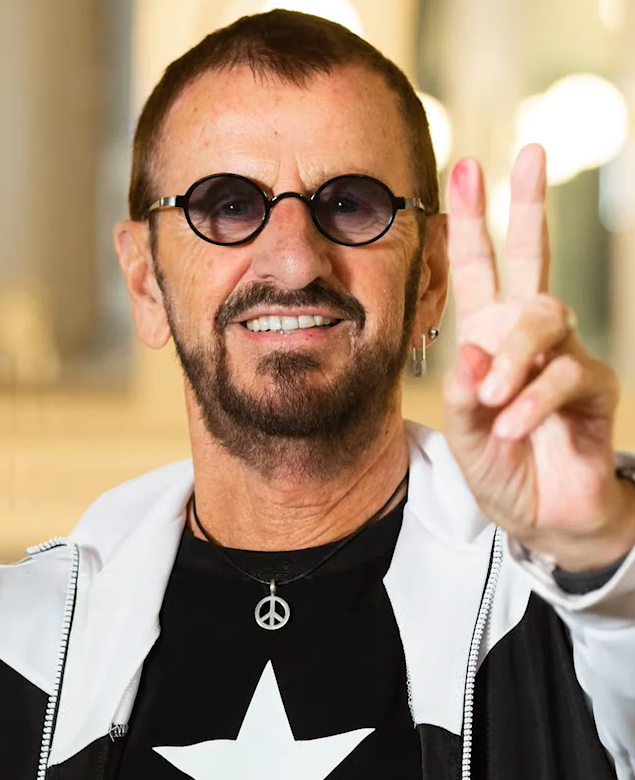 Ringo Starr mit kurzen Harren und Vollbart. Er trägt eine Sonnenbrille und zeigt das Peace-Zeichen.