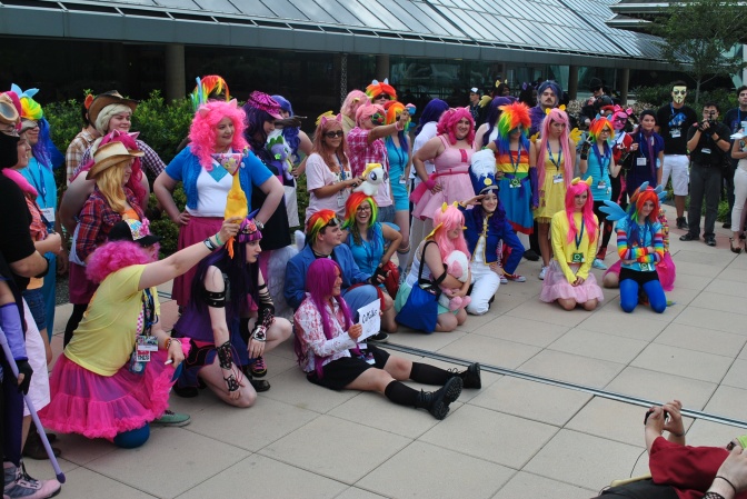 Eine große Gruppe erwachsener Menschen in bunten Plastikkostümen mit Perücken in Neonfarben. Sie haben sich fürs Foto nebeneinander aufgestellt und lächeln in die Kamera.
