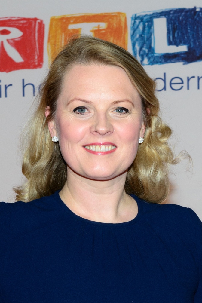 Patricia Kelly mit gelockten blonden Haaren in einem dunkelblauen Kleid. Sie steht lächelnd vor einer Wand mit dem RTL Logo.