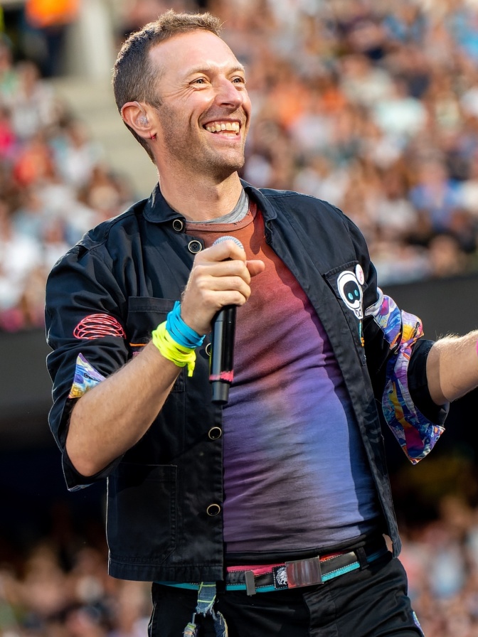 Chris Martin lächelt breit und hält ein Mikro in der Hand. Hinter ihm sieht man Publikum.