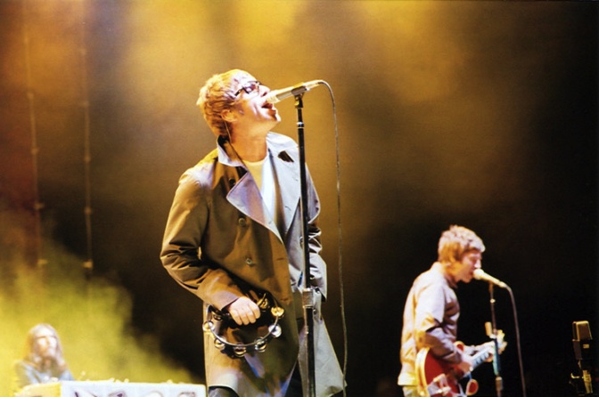 Liam und Noel Gallagher auf der Bühne. Sie singen in Mikros, die Bühne ist gelb beleuchtet.