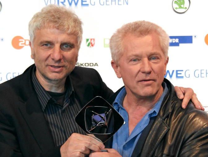 Udo Wachtveitl und Miroslav Nemec tragen beide schwarze Lederjacken und haben die Arme umeinander gelegt. Sie stehen vor einer Logowand.