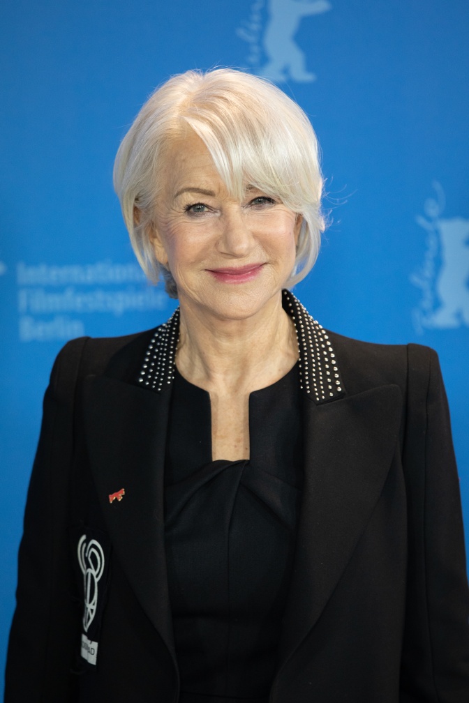 Helen Mirren mit weißen kinnlangen Haaren in einem schwarzen Kostüm. Sie lächelt leicht in die Kamera.