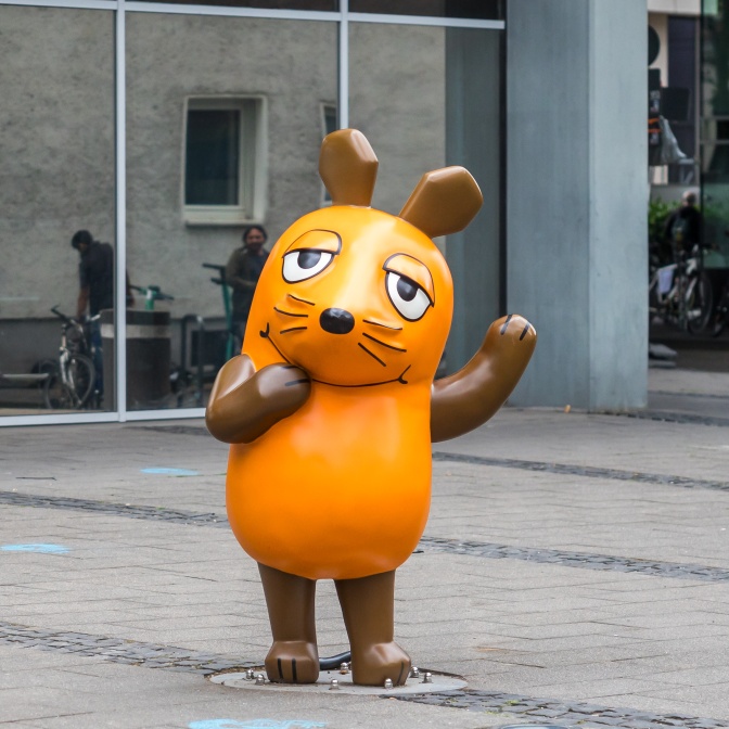 Eine Mausfigur in Braun und Orange. Sie hebt einen Arm und winkt.