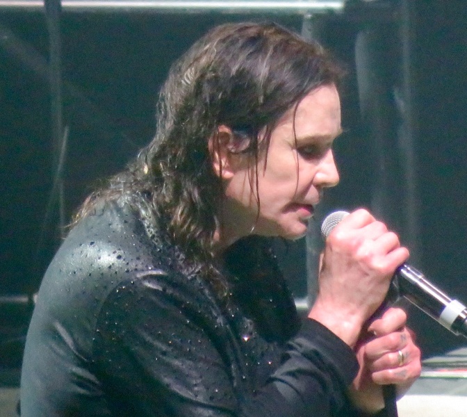 Ozzy Osbourne im Regen auf der Bühne. Er singt mit ernstem Blick in ein Mikrofon.