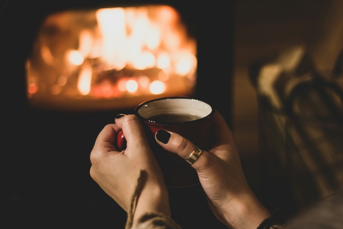 Eine Frau hält eine Tasse Kaffee in der Hand, im Hintergrund sieht man ein Kaminfeuer.