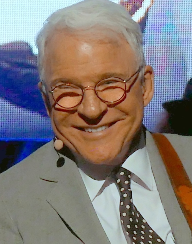 Steve Martin in Anzug und Krawatte. Er lächelt, seine Augen sind zum Teil von seiner Brille verdeckt.