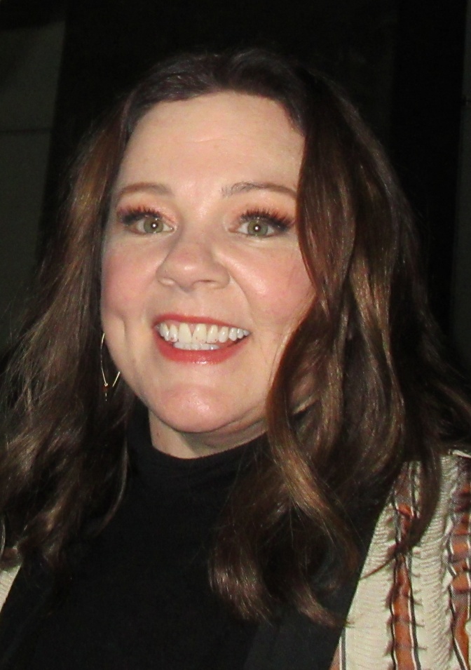 Melissa McCarthy mit gewellten langen braunen Haaren. Sie lächelt leicht an der Kamera vorbei.
