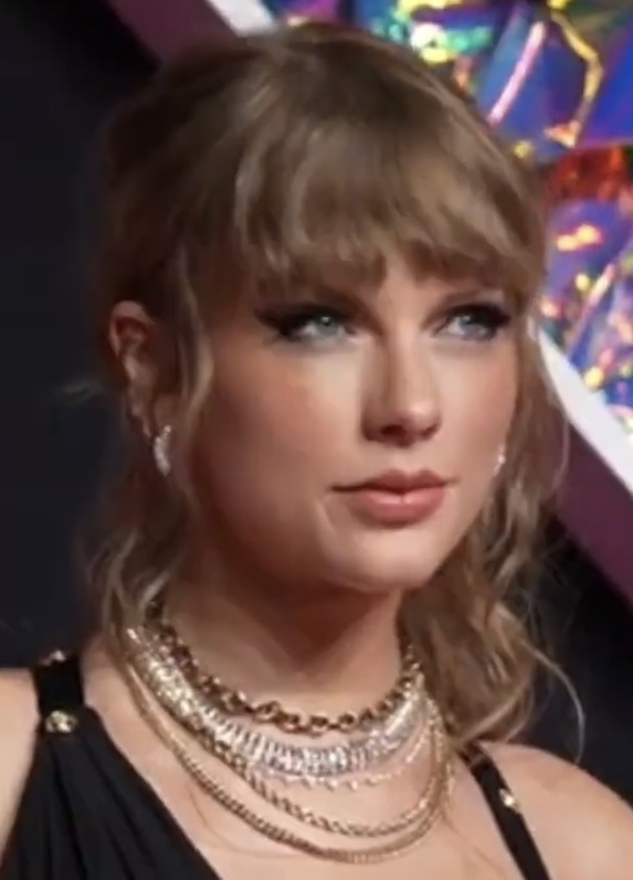 Taylor Swift mit zurückgesteckten. leicht gewellten Haaren. Sie trägt viel Schmuck und ein schwarzes Trägertop.