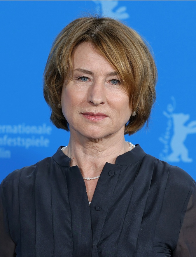 Corinna Harfouch in einer schwarzen Bluse vor einer blauen Wand. Sie schaut ernst in die Kamera.