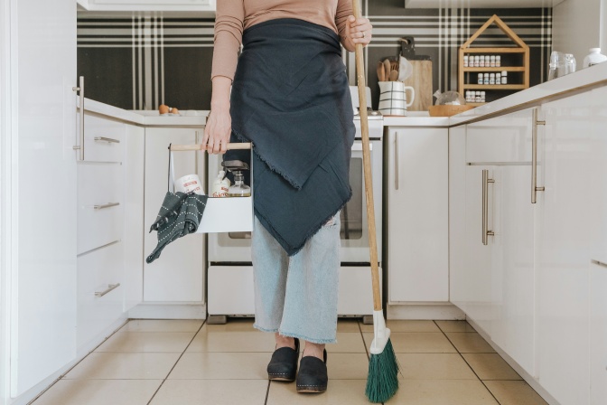 Eine Person mit Schürze steht in einer Küche. In einer Hand hält sie einen Besen, in der anderen einen Korb mit Reinigungsmitteln.