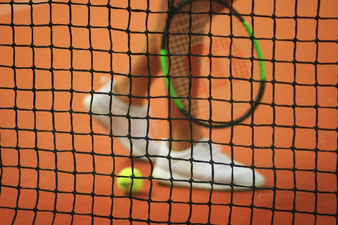 Ein Mann steht hinter einem Tennisnetz. Er hat einen Tennisschläger in der Hand, auf dem Boden liegt ein Tennisball.