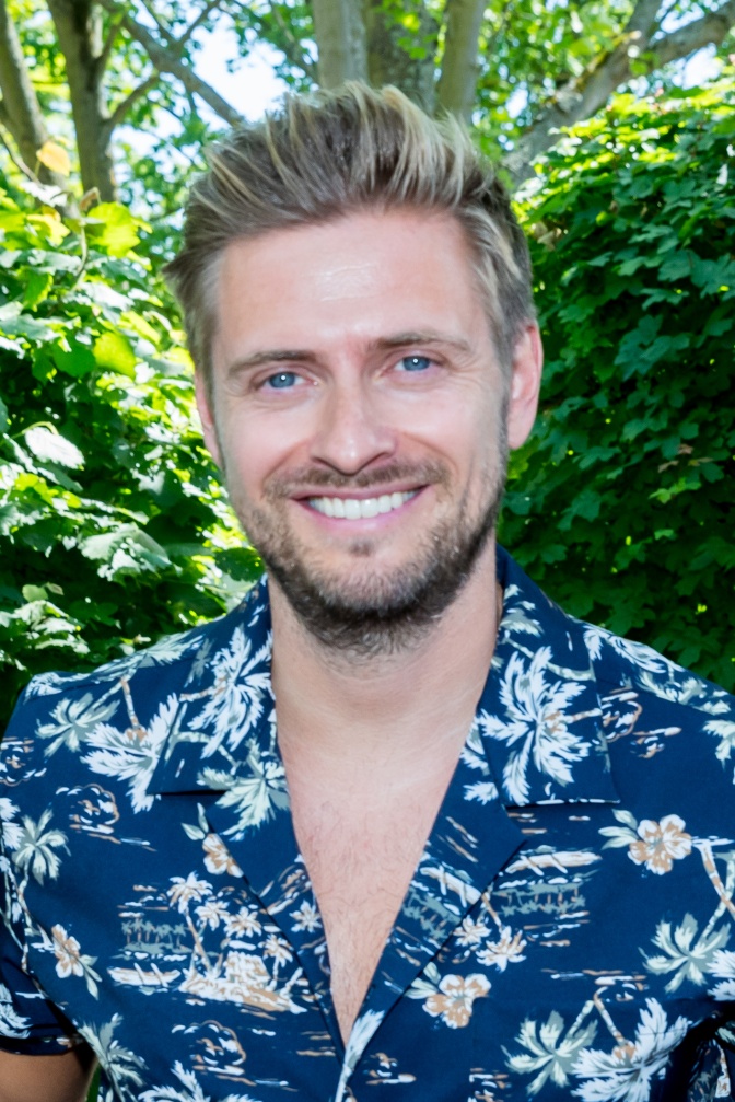 Jörn Schlönvoigt in einem blau-weißen Hawaiihemd. Er lächelt in die Kamera und steht im Freien.