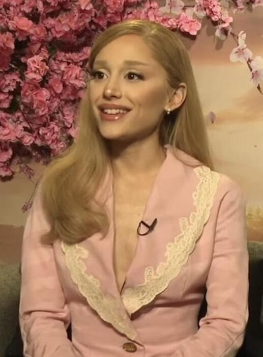 Ariana Grande in einem rosafarbenen Kostüm vor einer mit rosa Blumen bewachsenen Wand.