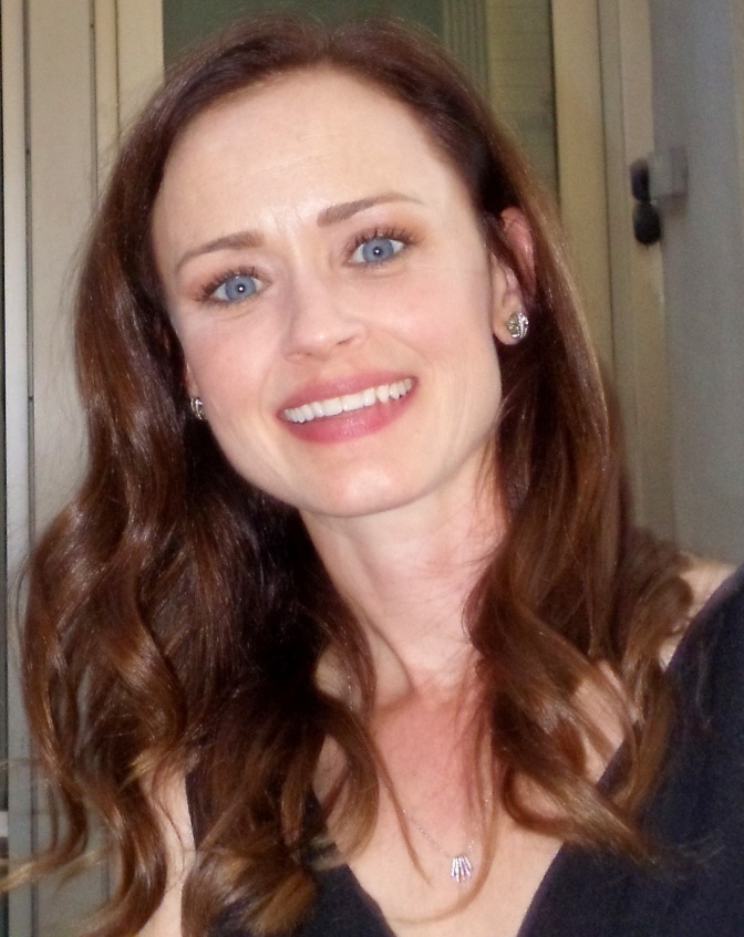 Alexis Bledel mit gewellten braunen Haaren in einem schwarzen Kleid. Sie lächelt direkt in die Kamera.