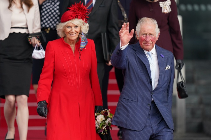König Charles und Königin Camilla gehen eine Treppe herunter. König Charles winkt.