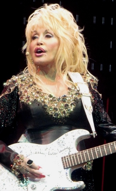 Dolly Parton in einem schwarzen Kleid mit Strass und Perlen am Kragen. Sie spielt eine weiße E-Gitarre und singt.
