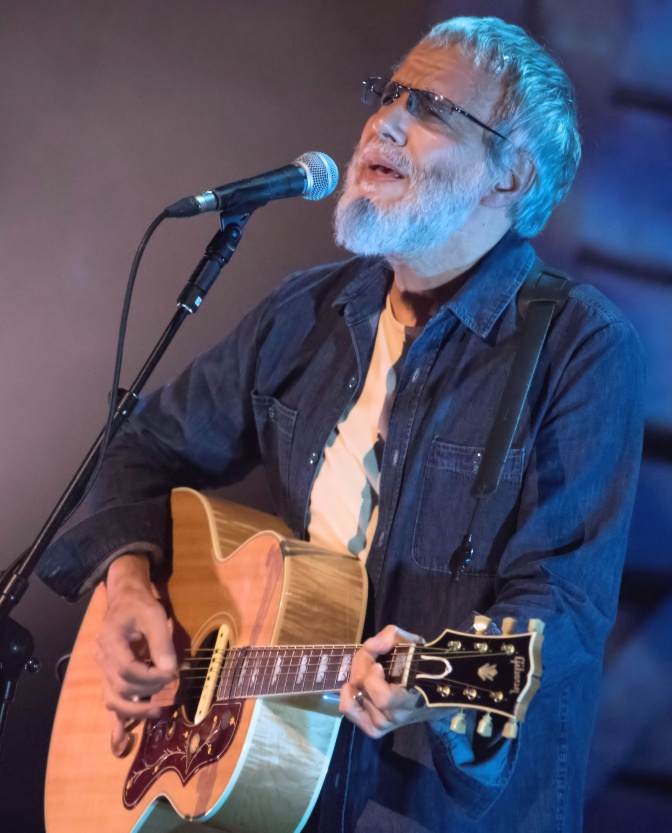 Cat Stevens mit weißen Haare, Vollbart und Brille. Er spielt Gitarre und singt dazu.