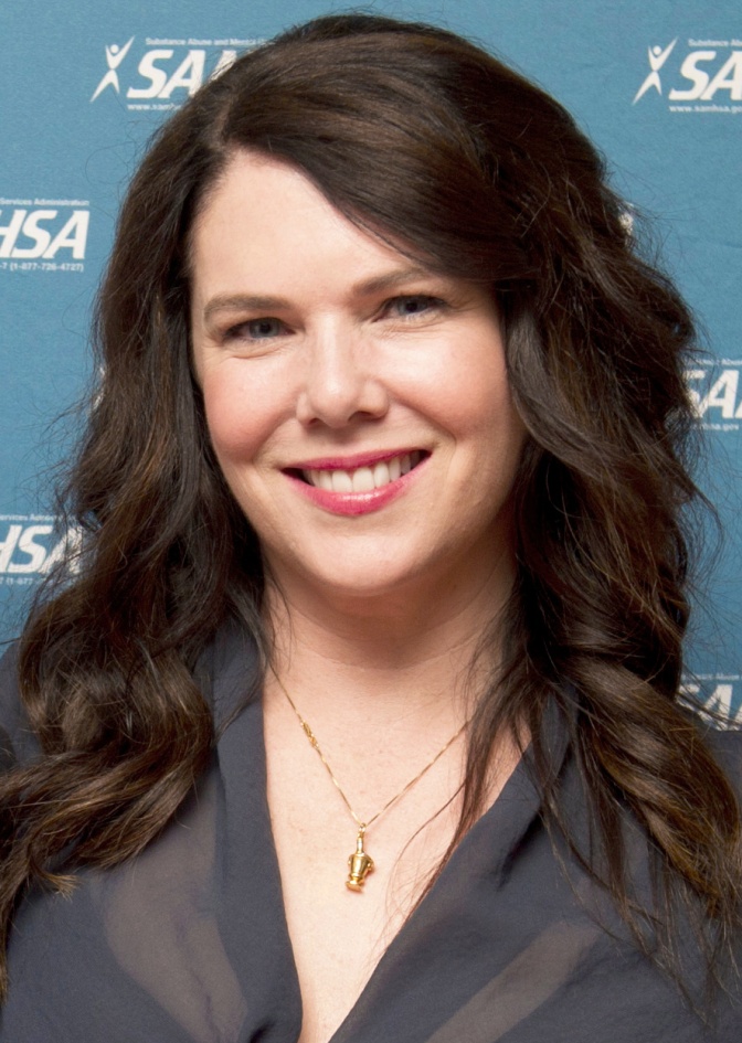 Lauren Graham mit gewellten braunen Haaren lächelnd vor einer Logowand.