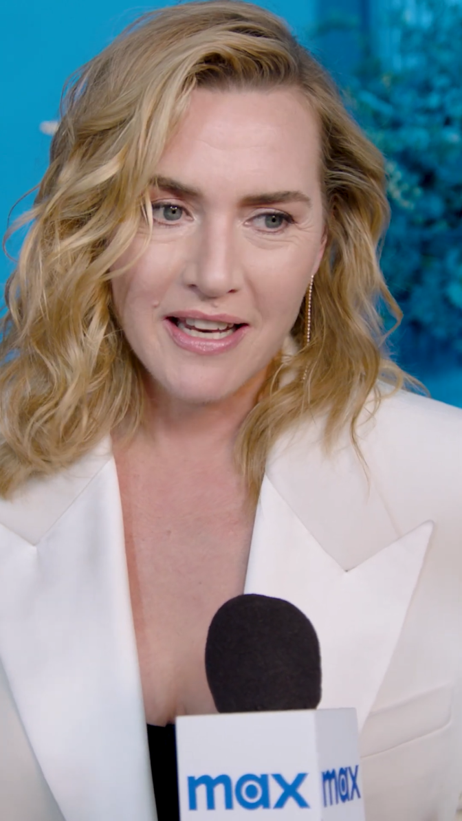 Kate Winslet in einem weißen Blazer mit gewellten blonden Haaren. Sie sitzt vor einem Mikrofon.