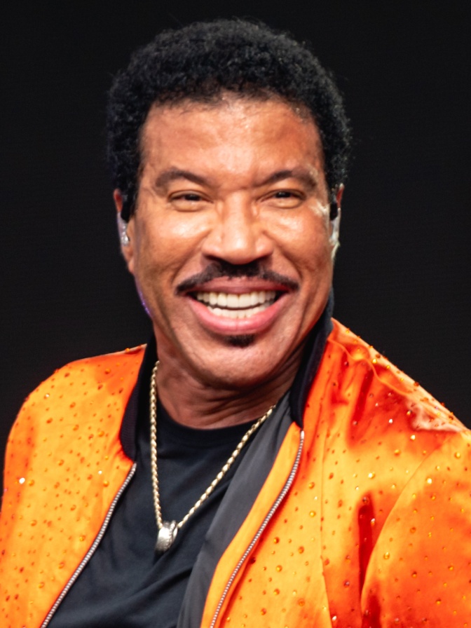 Lionel Richie mit Schnurrbart und Goldkette in einer leuchtend orangefarbenen Jacke mit Strass