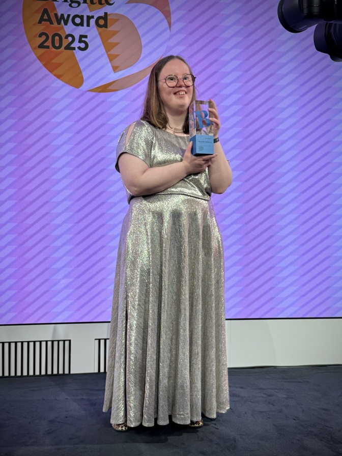 Natalie Dedreux steht in einem langen, silbernen Kleid auf der Bühne und hält einen Preis aus Plexiglas in der Hand. Sie lächelt. Hinter ihr sieht man auf eine rLeinwand das Logo des Brigitte Awards.