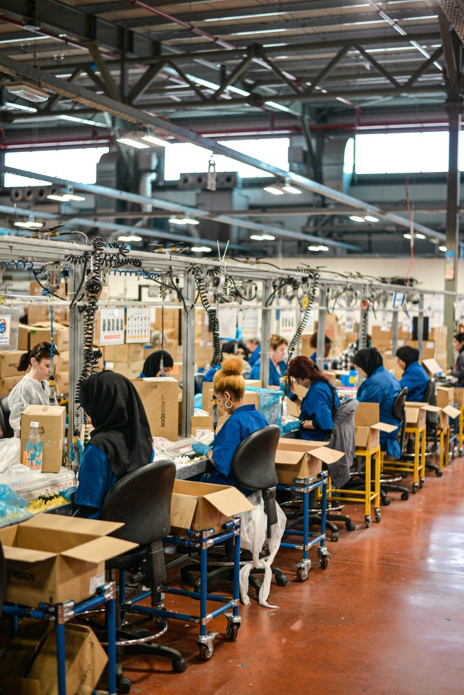 Frauen sitzen in einer Fabrik an Nähmaschinen. Einige von ihnen tragen Kopftücher.