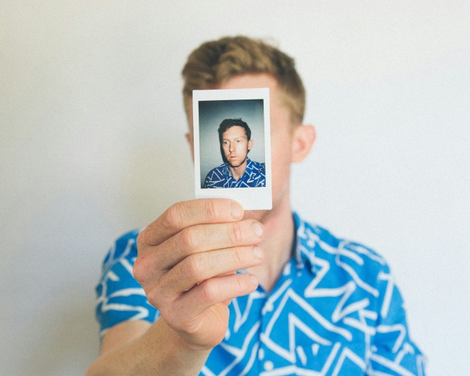 Ein Mann in einem blauen Hemd hält ein Polaroidfoto von sich selbst vor sein Gesicht.