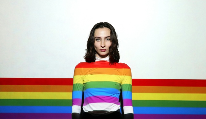 Eine Frau steht in einem weißen Pullover vor einer weißen Wand. Auf sie und die Wand wird ein Regenbogen projiziert.