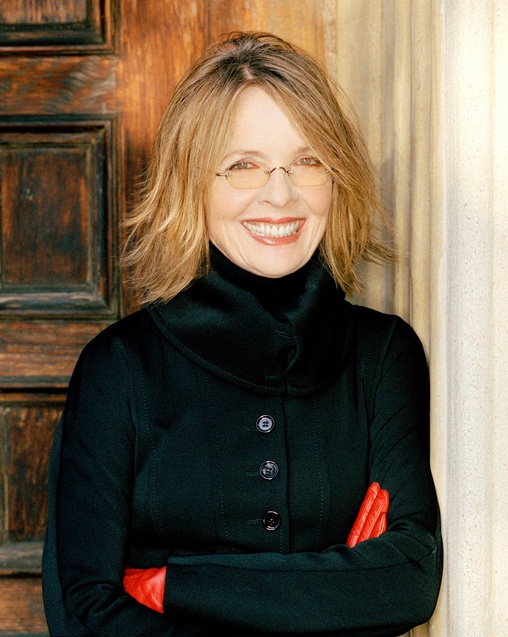 Diane Keaton in schwarzer Kleidung mit roten Handschuhen. Sie lehnt lächelnd an einem Türrahmen.