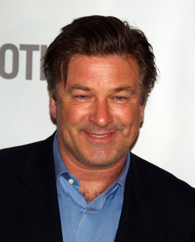 Alec Baldwin in einem Jeanshemd und schwarzem Sakko. Er steht lächelnd vor einer Logowand.