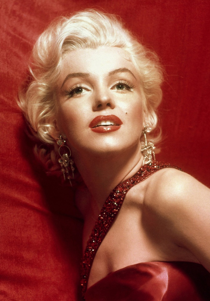 Marilyn Monroe mit in Wellen gelegten blonden Haaren in einem eleganten roten Kleid mit viel Schmuck.