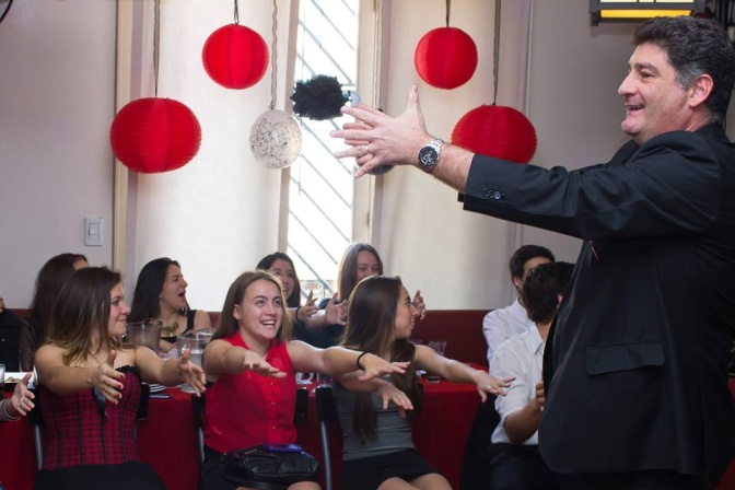 Ein Mann in Anzug steht mit ausgebreiteten Armen vor dem Publikum. Im Hintergrund hängen Lampions in rot und schwarz.