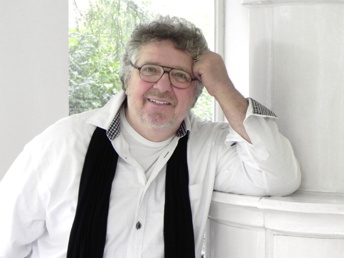 Michael Schanze lehnt an der Wand und hat den Kopf auf die Hand gestützt. Er schaut direkt in die Kamera und lächelt.