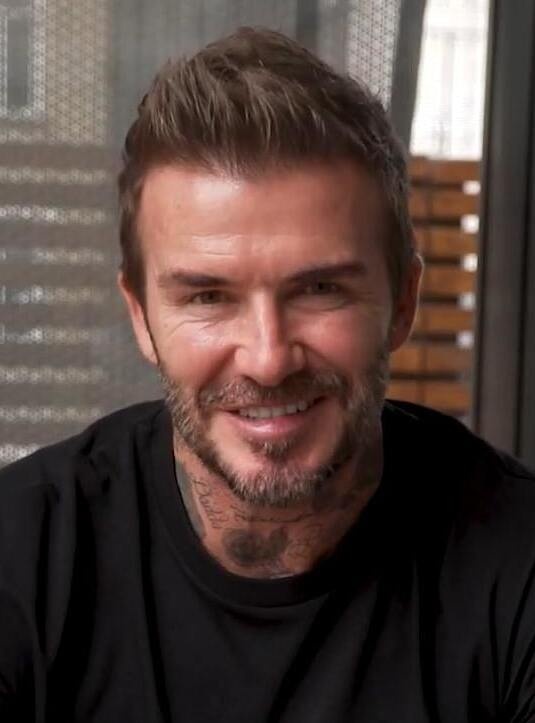 David Beckham mit Vollbart und Tattoo am Hals. Er schaut lächelnd direkt in die Kamera.