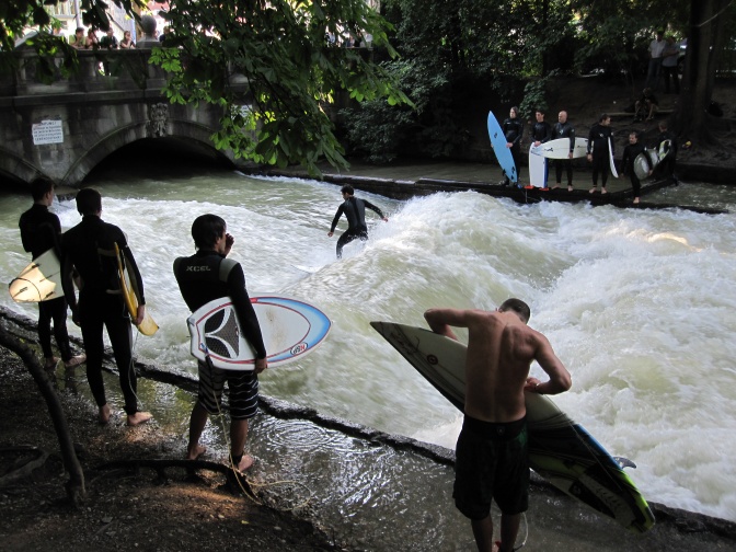 Ein Surfer surft auf der Welle im Bach, mehrere andere stehen in Neoprenanzügen mit ihren Surfbrettern wartend daneben.