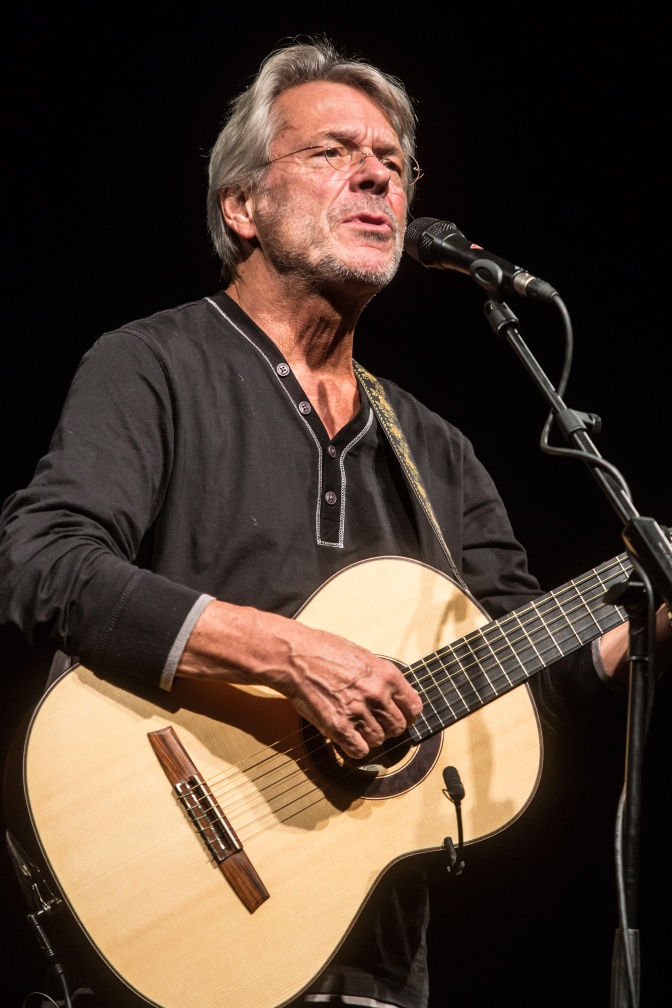 Reinhard Mey spielt eine akustische Gitarre und singt. Er hat graue Haare, einen Dreitagebart und trägt Brille.