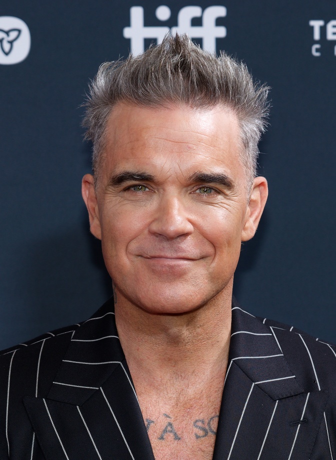 Robbie Williams mit grauen Haaren in einem Sakko mit Nadelstreifen. Er steht vor einer Logowand und lächelt leicht in die Kamera.