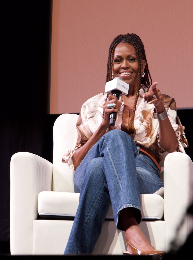 Michelle Obama mit Braids. Sie sitzt in einem weißen Sessel und spricht in ein Mikrofon.
