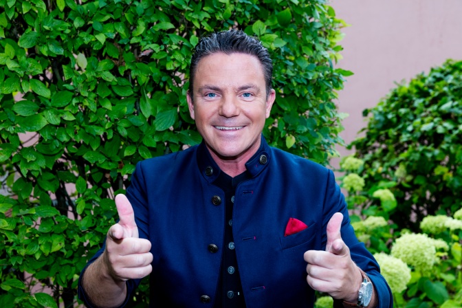 Stefan Mross in einem lilafarbenen Sakko vor einer Bühnendekoration aus künstlichen Büschen. Er streckt lächelnd beide Daumen in die Höhe.