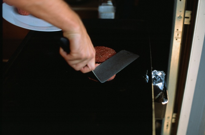 Eine Person drückt ein Burgerpatty beim Braten mit einer Kelle platt.