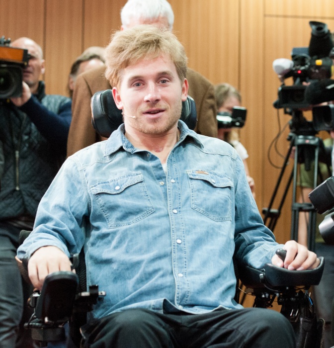 Samuel Koch trägt ein Jeanshemd und sitzt in seinem Elektrorollstuhl. Er schaut direkt in die Kamera.